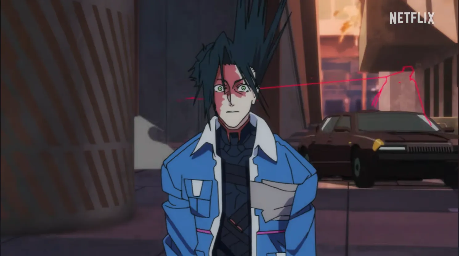 A man standing with blue coat and black hair. A car rounds the corner behind him, trailing red lightning